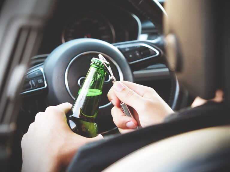 close up of person opening a bottle of beer while sitting in front of a steering wheel of a car